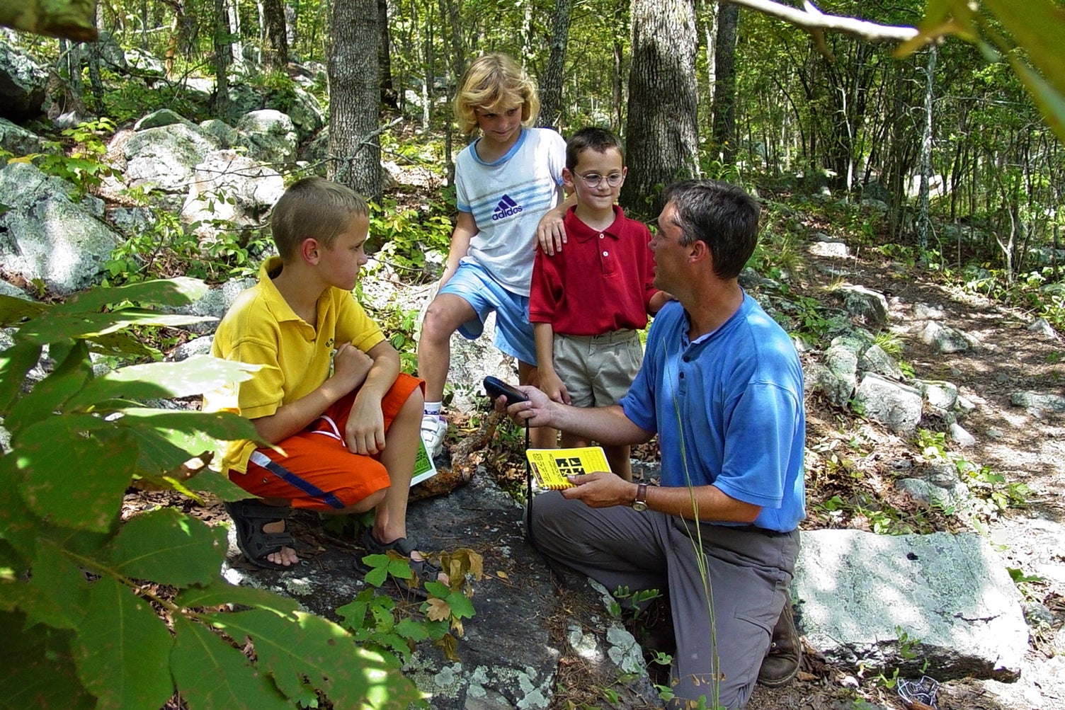 an adult and three children trailside, discussing geocaching. The adult holds a GPS device.